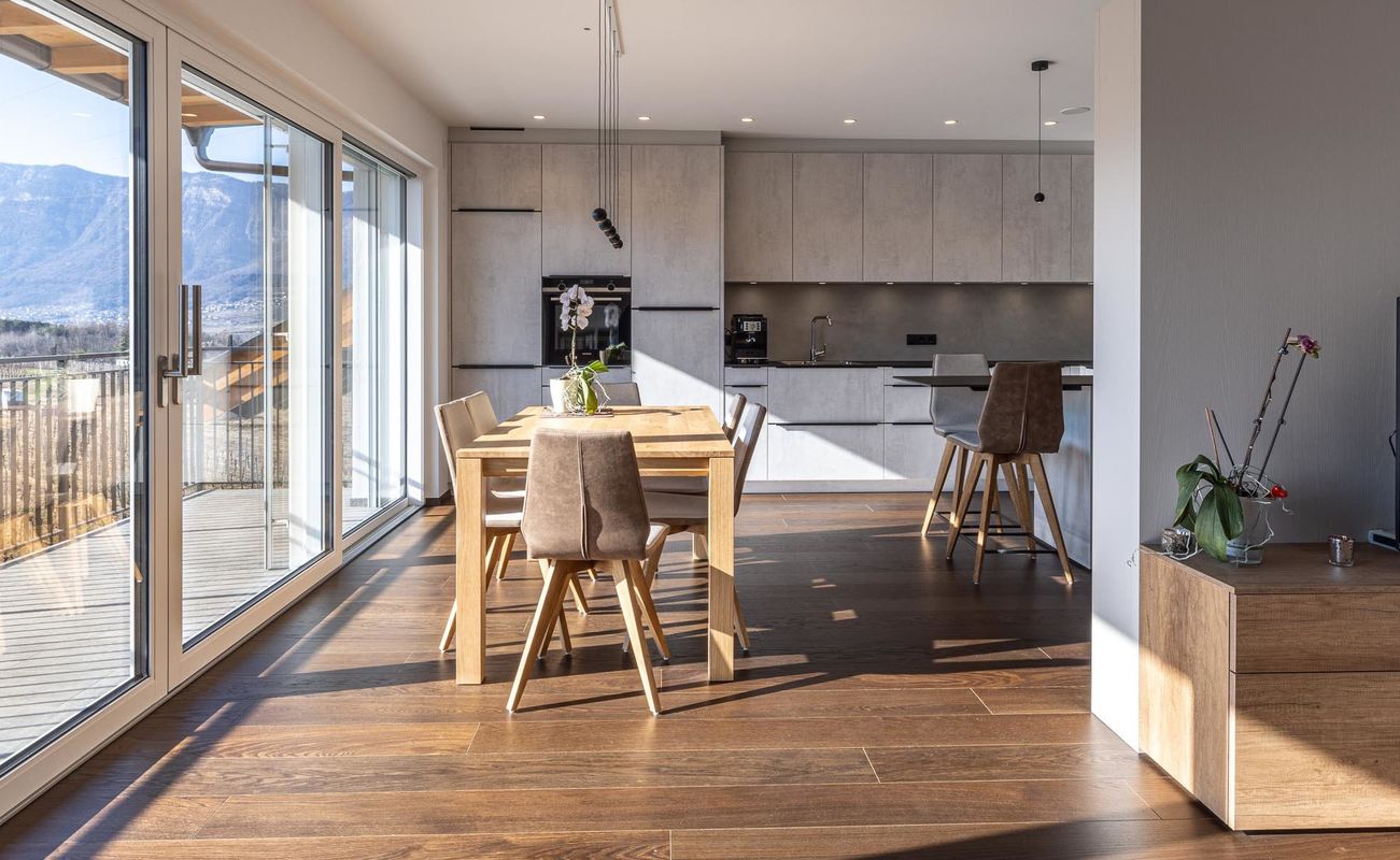Moderne Wohnung mit offener Küche und Essbereich, großen Fenstern und Blick auf die Berge. Helle Holzoberflächen und minimalistische Einrichtung.