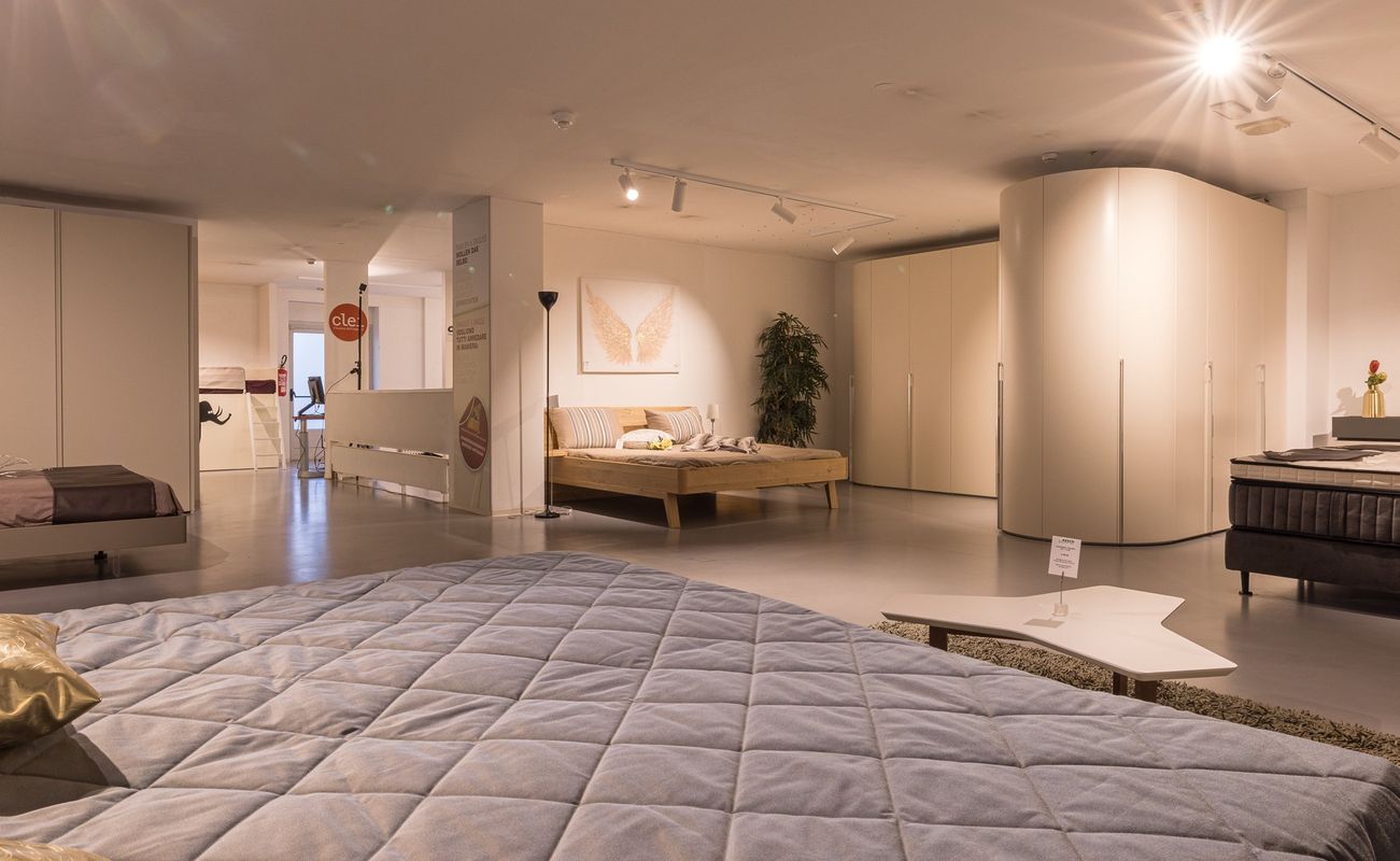 Ein modernes Schlafzimmer-Showroom mit mehreren Betten, Schränken und einem Teppich. Helle Beleuchtung und neutrale Farben dominieren den Raum.