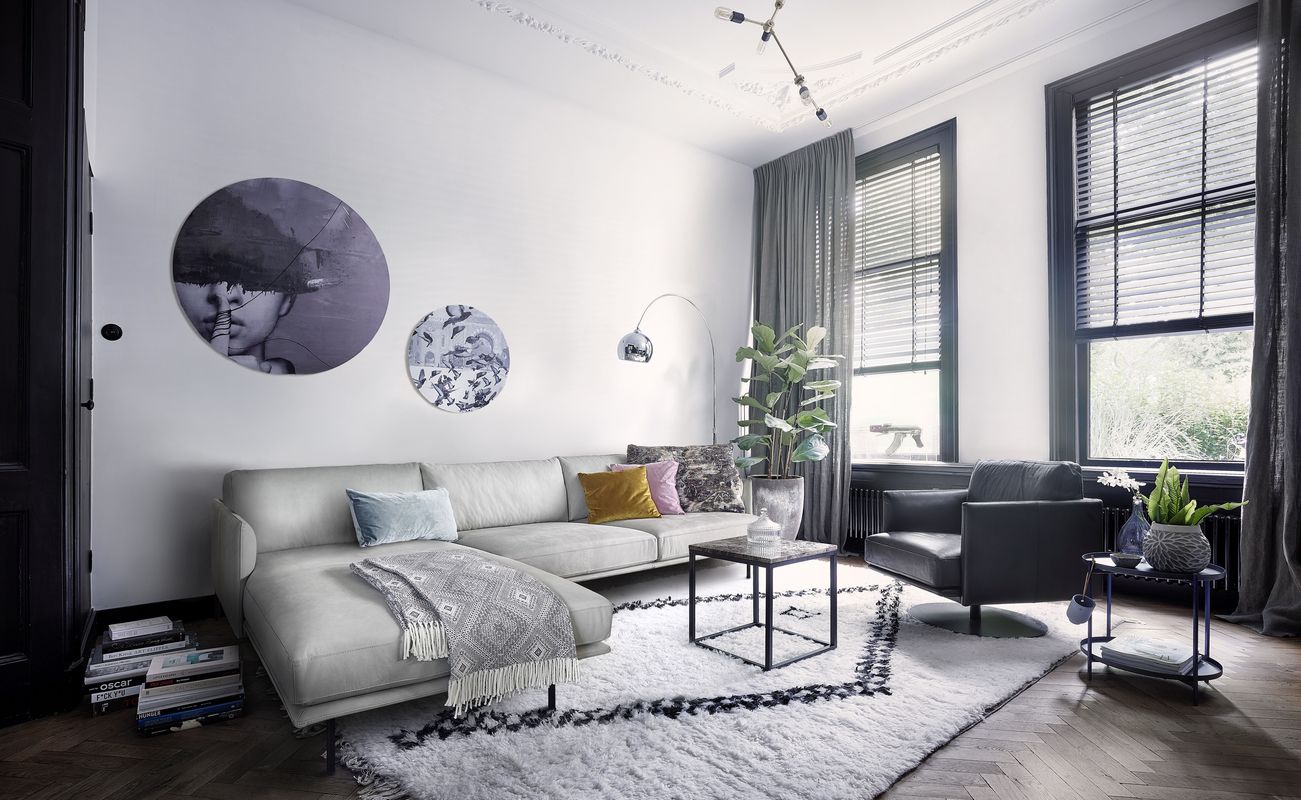 Modernes Wohnzimmer mit einem weißen Sofa, einem schwarzen Sessel, einem Teppich und Kunstwerken an der Wand. Fenster mit Vorhängen und Pflanzen.