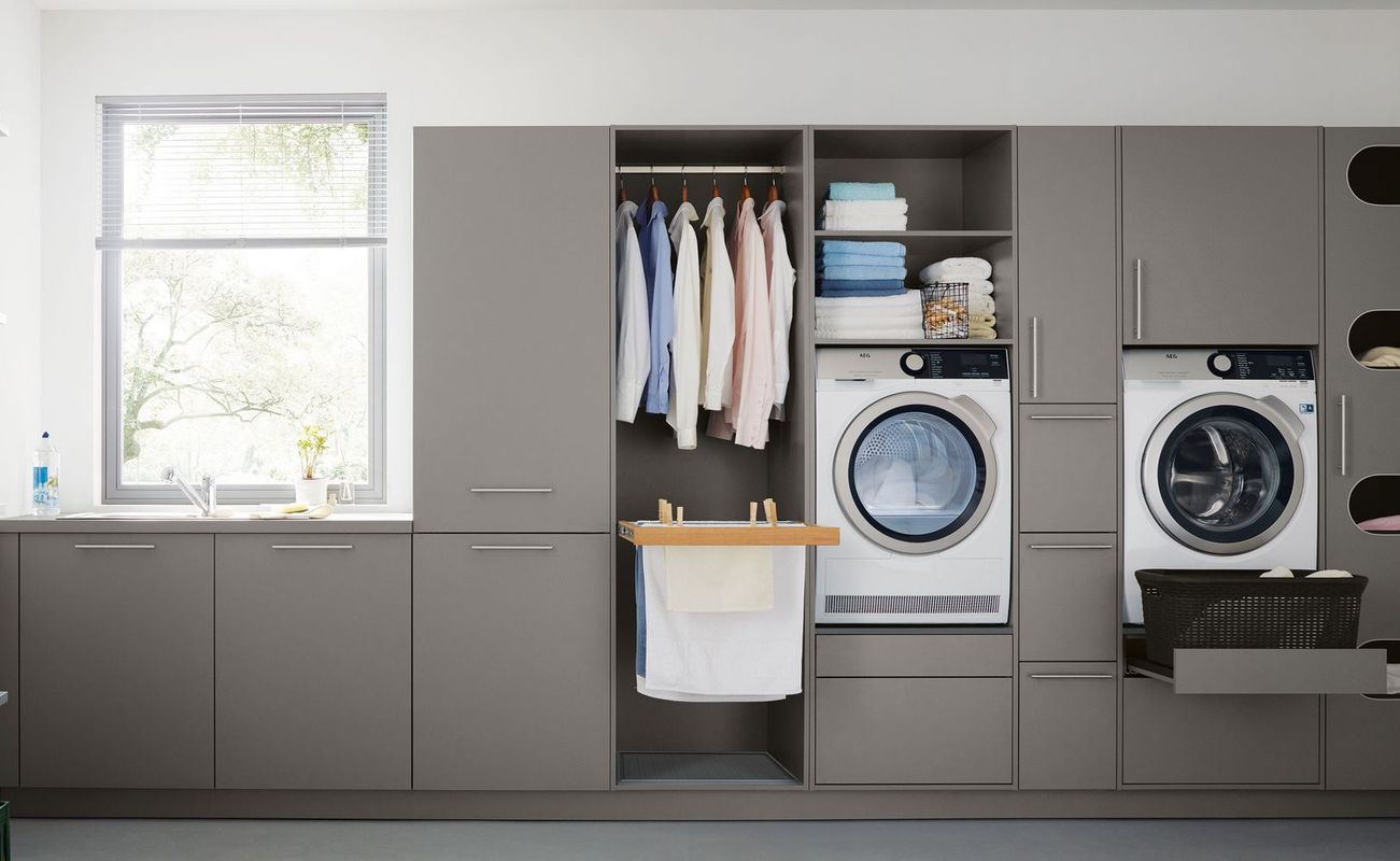 Moderne Waschküche mit grauen Schränken, Waschmaschine, Trockner und aufgeräumten Regalen. Kleidung und Handtücher sind ordentlich aufgehängt.